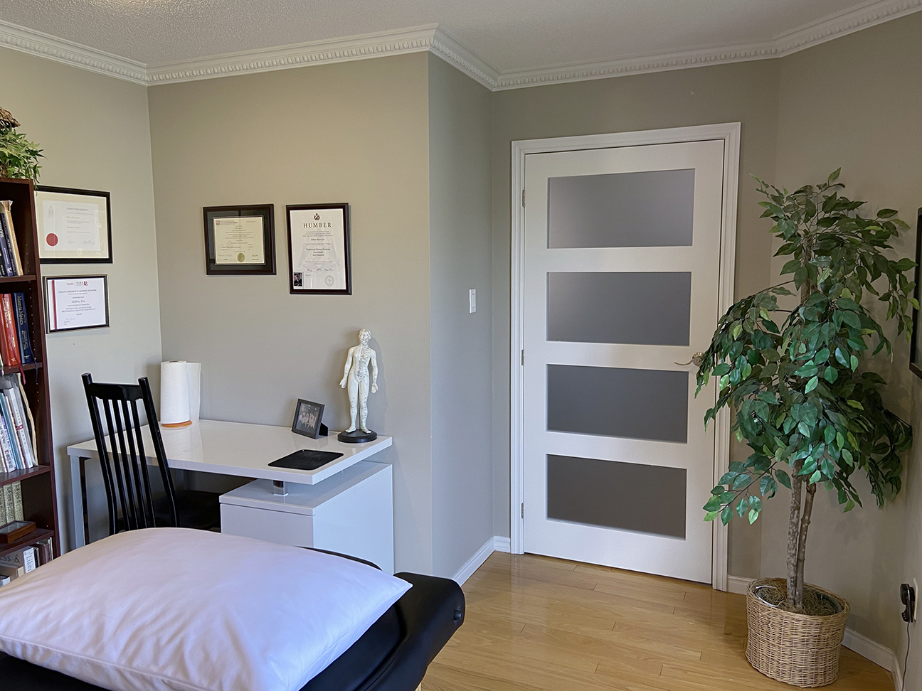 photograph showing reverse view of clinic across massage table toward a desk with diplomas and certificates in frames on the walls, the clinic door closed, and a tall green plant in the corner